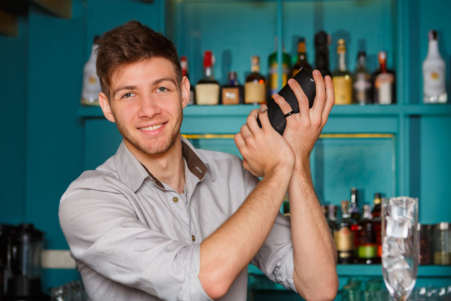 Young tattooed male bartender  prepares cocktail using steel shaker behind bar counter
